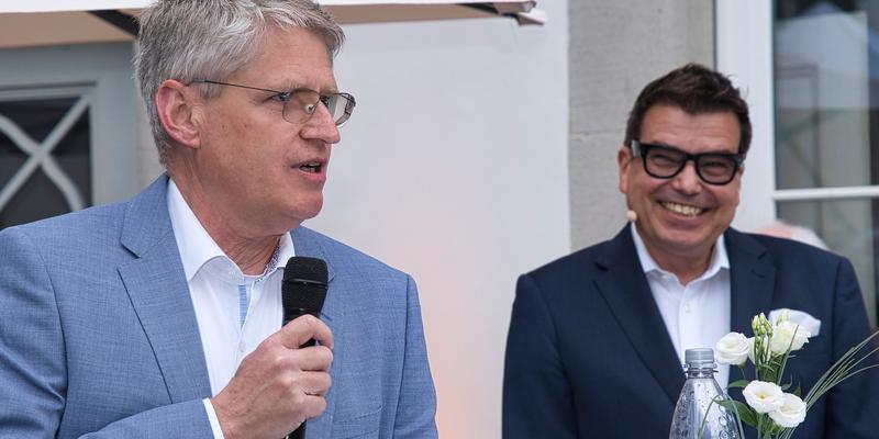 A man is talking holding a microphone, another man is smiling in the background.