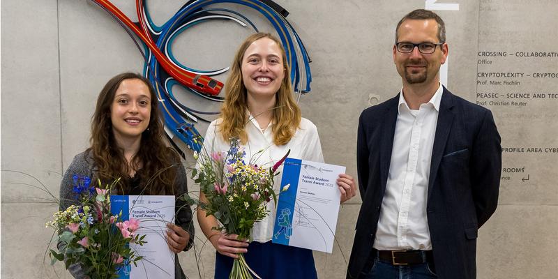 Two Women with certificates and flowers and a man in front of a piece of artwork