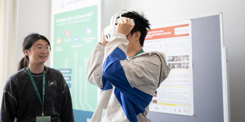 Zwei Personen vor einer Stellwand mit einem Poster; eine Person trägt eine VR-Brille, die andere Person schaut sie an.