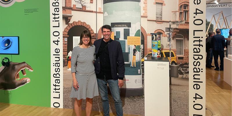 Two people are standing arm in arm next to a small model of the Litfaßsäule 4.0 and in front of a large photograph of the Litfaßsäule 4.0