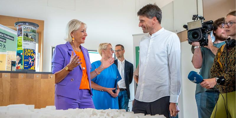Eine bunt angezogene Frau steht vor einem großen Stadtmodell und unterhält sich mit einem Mann; links steht das Modell einer Litfaßsäule mit digitalem Bildschirm im Kapitell und im Hintergrund sind Menschen mit Kameras und weitere Forschende zu sehen.