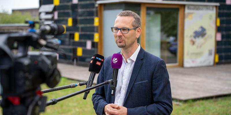 A man stands in front of the eHUB with two mikrophones in front of him and a camera