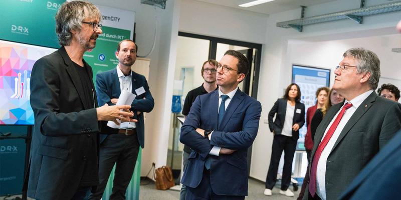 Eine Person spricht und drei weitere hören interessiert zu. Im Hintergrund stehen weitere Personen, die auf den Sprecher blicken.