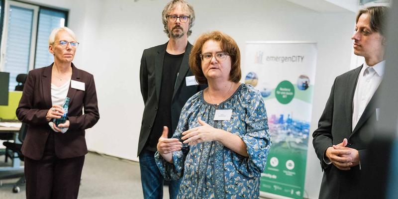 Eine Frau steht in der Mitte des Bildes und gestikuliert beim Sprechen. Um sie herum stehen drei weitere Personen und im Hintergrund ein Roll-Up mit dem emergenCITY Logo.