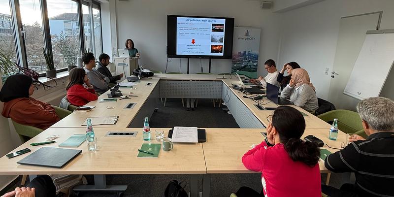 Mehrere Personen sitzen um Tische herum und hören einer Person zu, die vorne eine Präsentation hält.