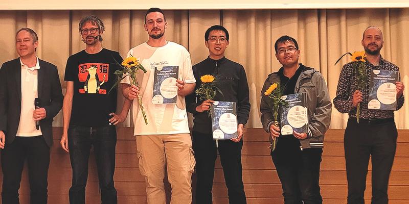 Six men stand side by side on a stage. Four of them are holding the award and a flower in their hands.