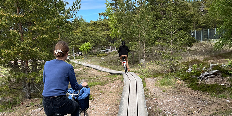 Two people on bicycles can be seen from behind on a narrow path. There is a forest around them.