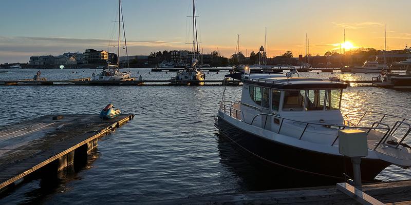 A small ship is moored at a jetty. Blue water stretches out behind it, reflecting the sunset. Further back, more ships and parts of the city can be seen.