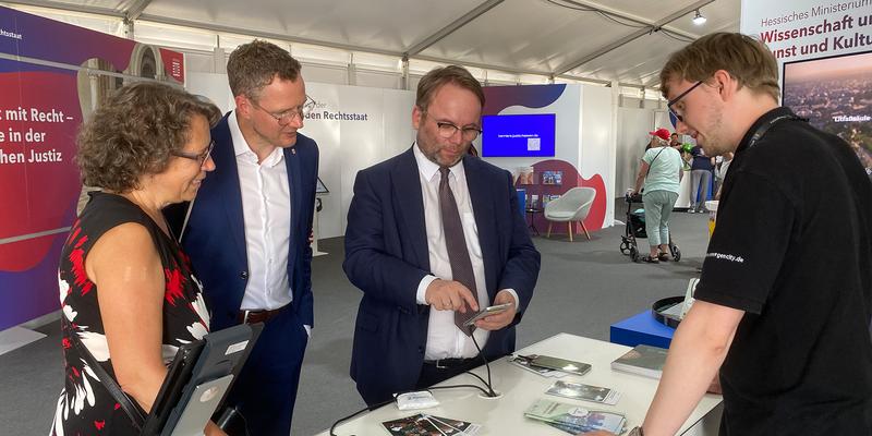 Four people are standing at a stand in a trade fair tent, one person, Science Minister Timon Gremmels, is trying out a smartphone game.