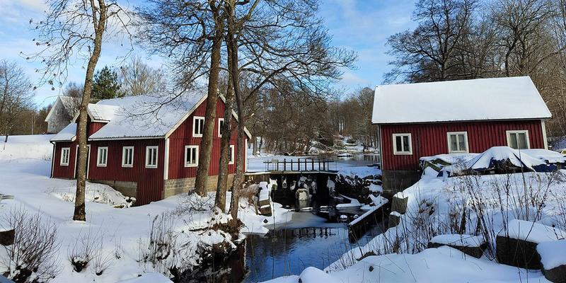 Two red houses stand next to a stream. In the background is a forest; everything is covered in snow.
