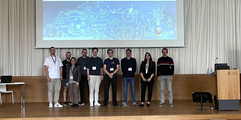 Eine Gruppe von Personen steht auf einer Bühne vor dem Titelbild der Präsentation